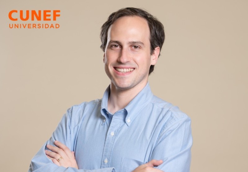 Un hombre sonriente con camisa azul se encuentra frente a un fondo beige, mostrando confianza.