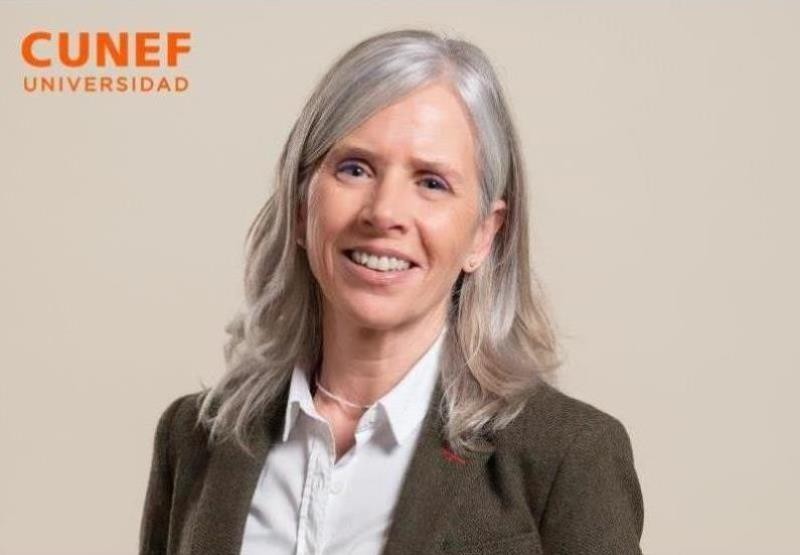 La imagen muestra a una mujer sonriente con cabello largo y canoso, vestida con una chaqueta oscura frente a un fondo beige.